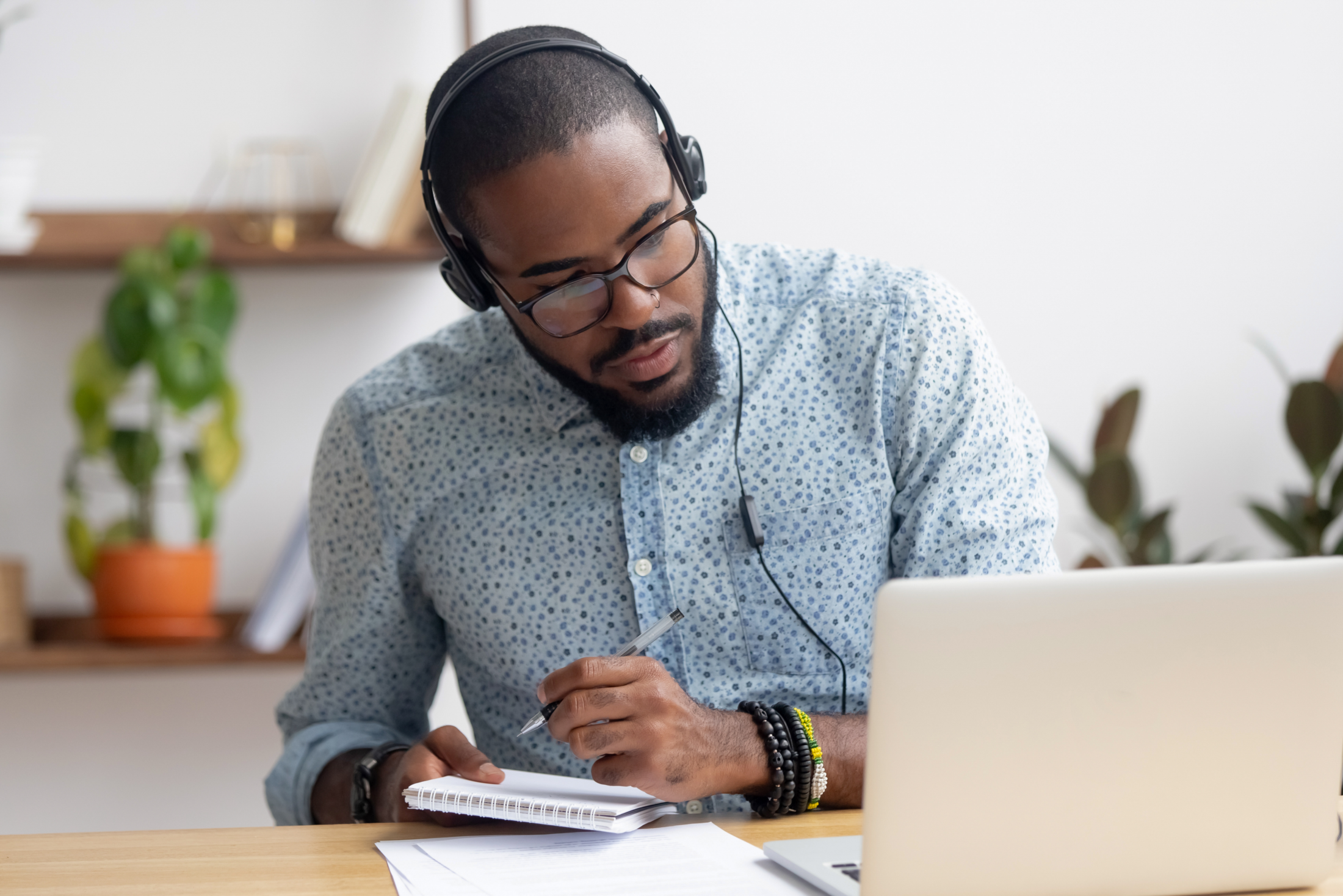 Person wearing headphones takes notes while viewing a laptop.